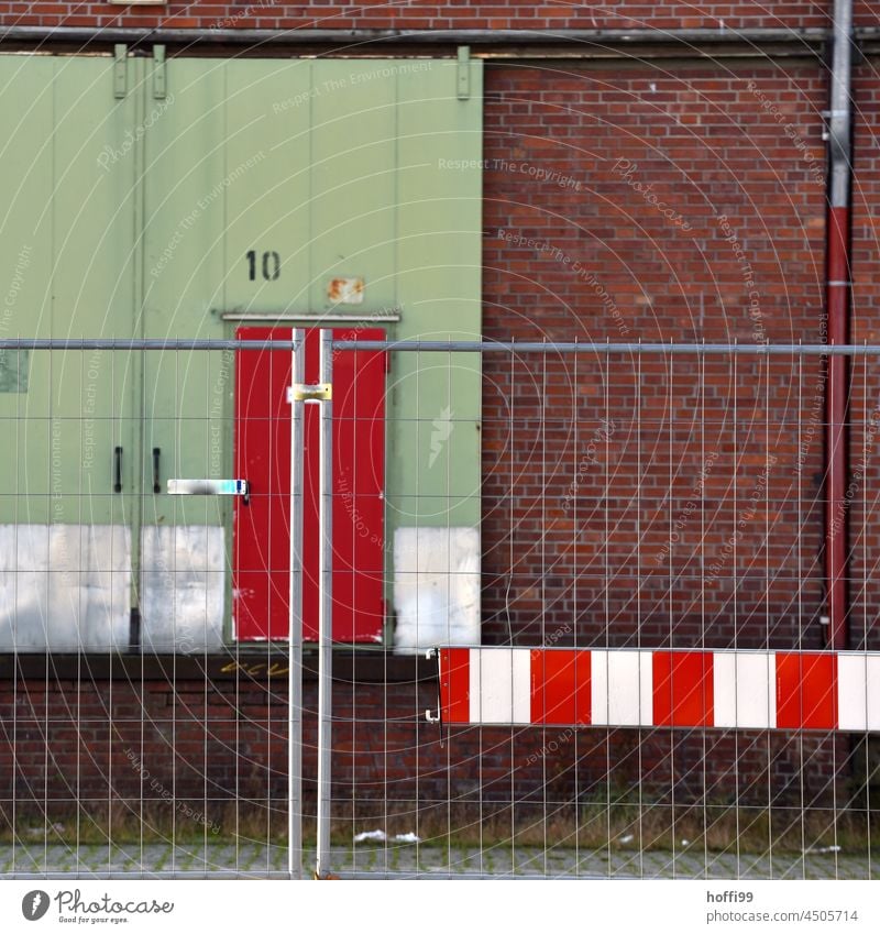 Warning sign on construction fence in front of rolling gate with red door on warehouse with downpipe and red bricks 10 cordon White Red Barque Barrier Hoarding