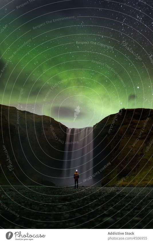 Anonymous traveler observing starry sky with aurora borealis near picturesque waterfall person admire night polar cliff nature landscape light tourist scenic