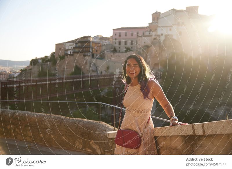 Cheerful ethnic woman in dress leaning on stone border sunset historic town cityscape hill attract happy enjoy female fence footbridge old positive aged travel