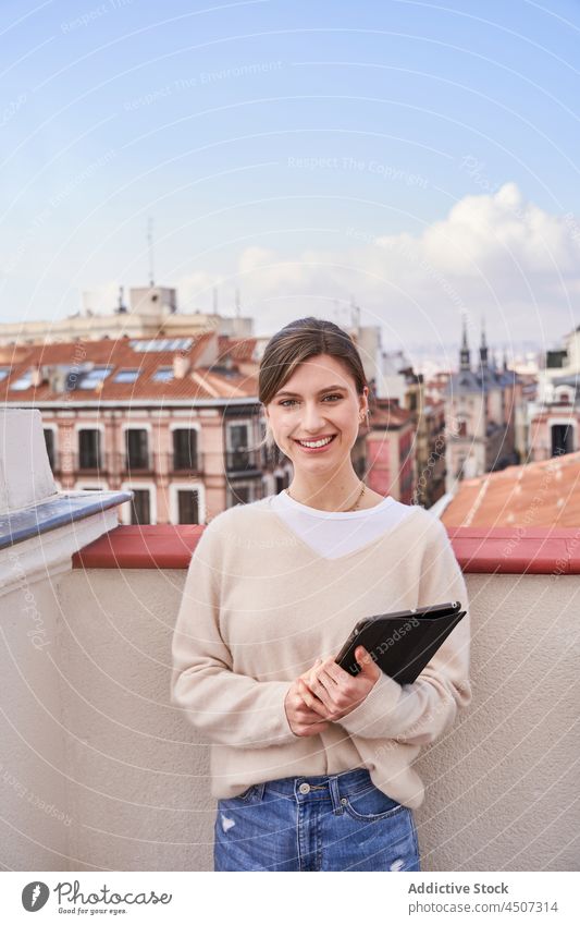 Smiling woman with tablet standing on balcony using internet appearance rooftop positive online charming female phone terrace casual jeans optimist
