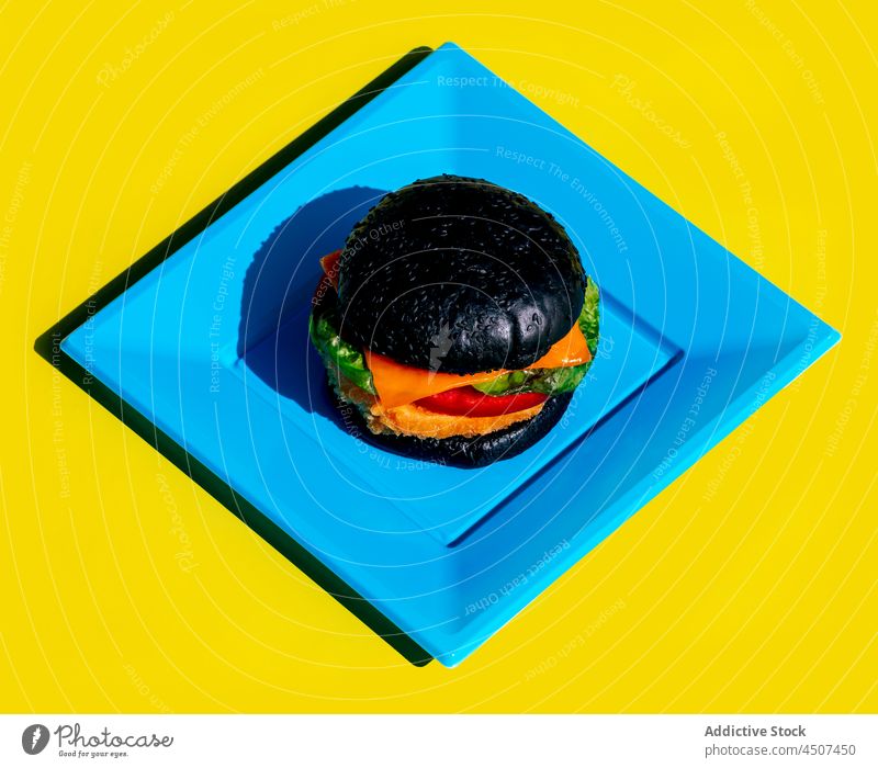 Black burger on square blue plate junk food colorful bright geometry dish lunch portion design simple minimal shape fast food bun vegetable multicolored meal