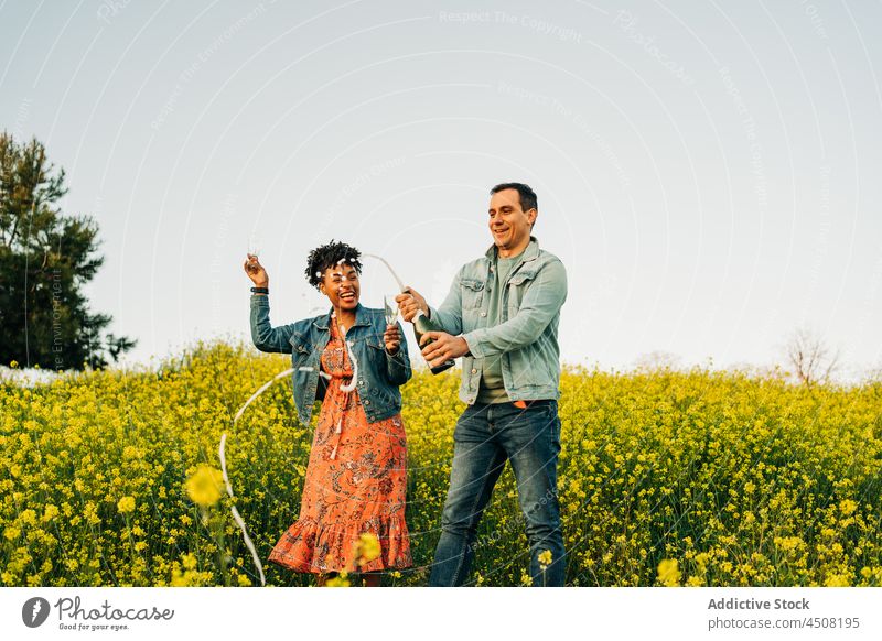 Cheerful young multiracial couple opening champagne bottle in meadow cheerful celebrate date romantic love relationship together happy countryside boyfriend
