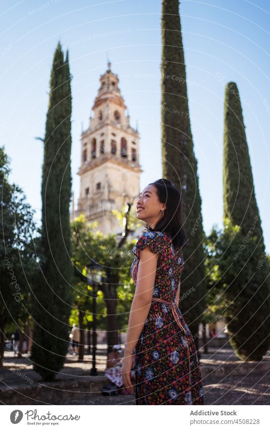 Positive Asian woman near historic building and trees street town aged tourist architecture observe medieval plant admire flora ancient colorful classic culture