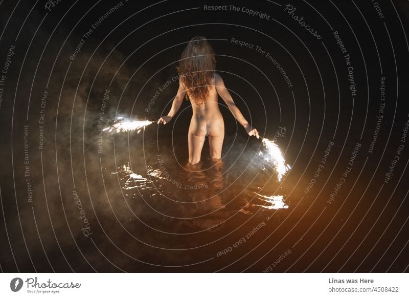 A wild and naked girl is swimming at night. Erotic image of a brunette model. Fireworks in her hands, sexy back show her perfect curves, smoke, and dark water makes this scene moody and sensual.