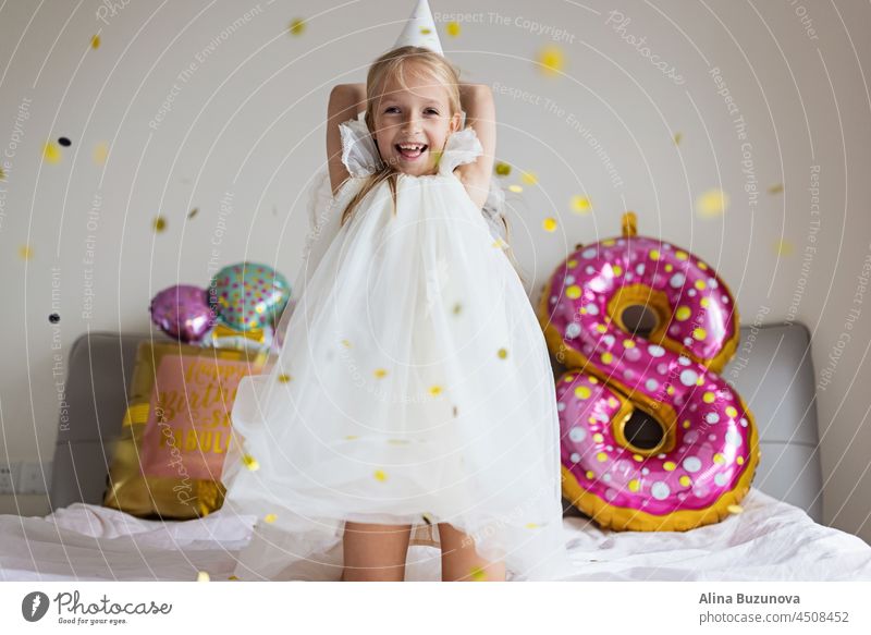 Cite little caucasian girl celebrating eight years old birthday at home. Stylish Kid wearing fashionable dress and having fun with confetti on bed Confetti
