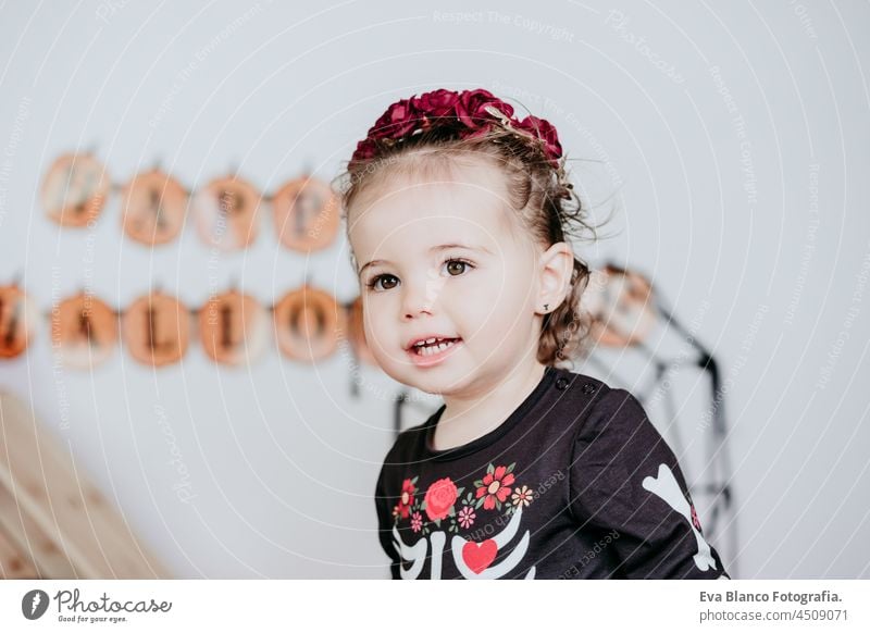 close up of beautiful one year old caucasian girl in halloween costume at home with Halloween decoration, holding balloon. Lifestyle indoors. Halloween party concept.