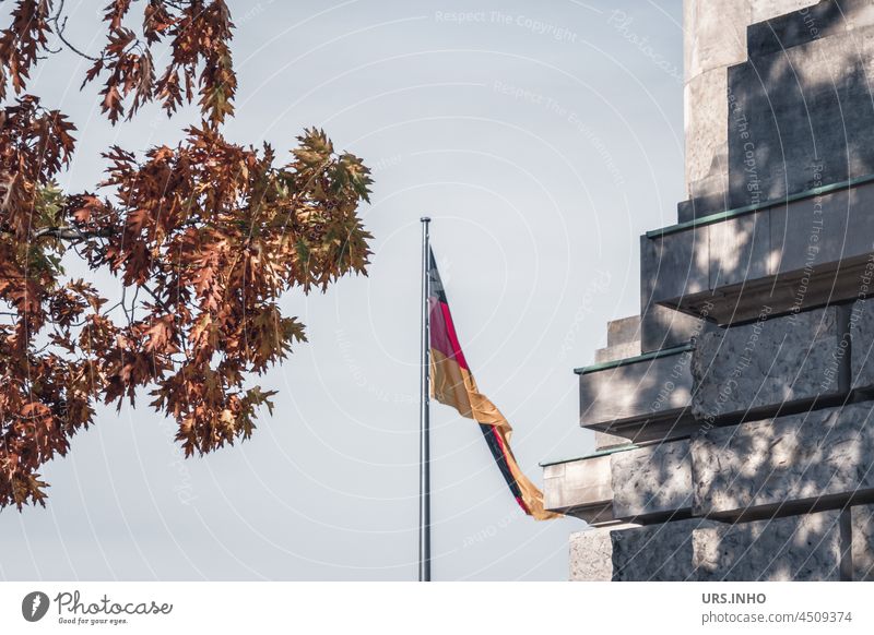 a German flag is flying on a pole between the tree top in the colours of autumn and the shadow of its autumnal leaves on the stones of the building Germany