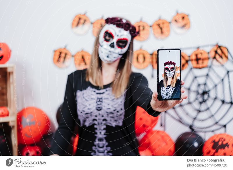 woman wearing mexican face mask during halloween celebration. taking selfie with mobile phone. woman wearing skeleton costume and red roses diadem on head. Halloween party concept