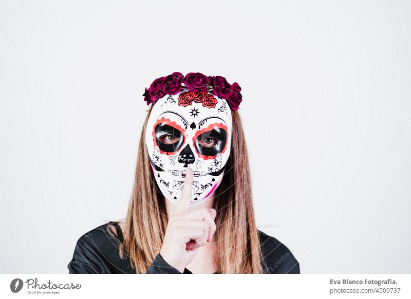 woman wearing mexican face mask during halloween celebration. skeleton costume and red roses diadem on head. woman making silence gesture with finger. Halloween party concept