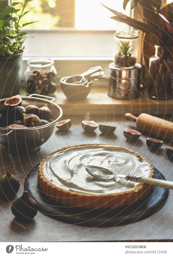 Cake with creme and spoon on kitchen table with figs in sieve, rolling pin and baking equipment at window background. Baking preparation at home with fresh ingredients. Front view.