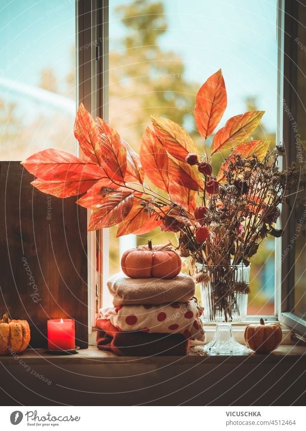 Cozy autumn still life with candle, sweaters, pumpkin and autumn branches on window sill background. Hygge lifestyle at home with warm clothes and cozy decoration. Front view.