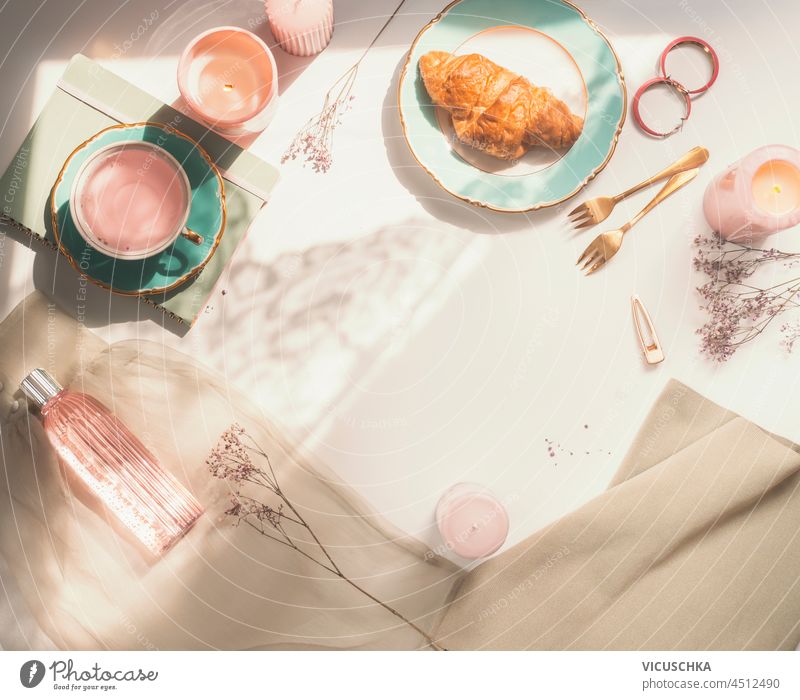 Modern morning routine with croissant, flowers tea, scented candles, forks, pink perfume and notebook on white table. Romantic feminine background. Top view with copy space.