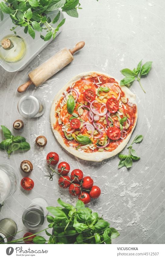 Homemade pizza with onion, salami, tomatoes and basil on grey kitchen table with wooden rolling pin, mushrooms and kitchen utensils. Preparing italian food at home with fresh ingredients. Top view.
