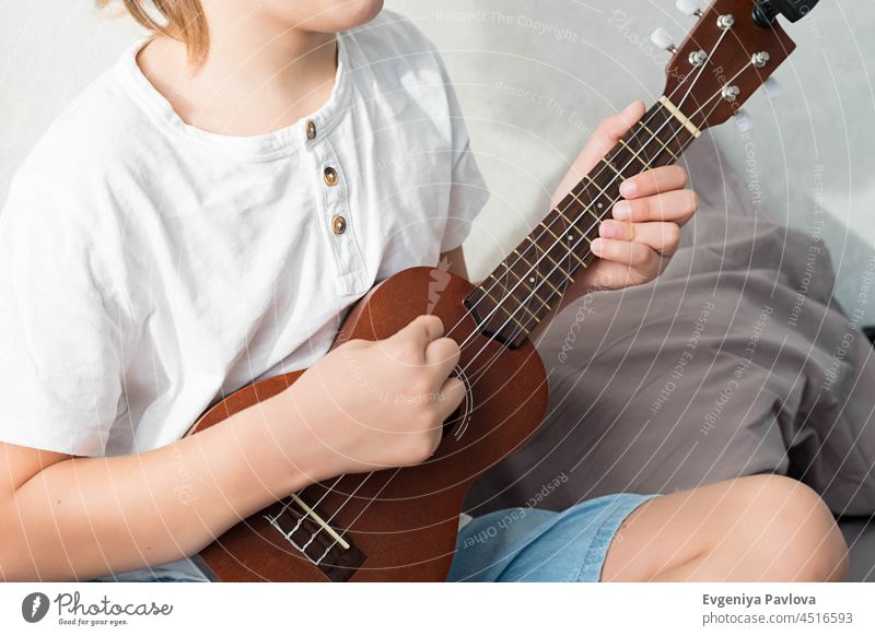 Young boy tuning ukulele at home. Blond haired boy sitting on couch playing acoustic guitar. young cheerful hobby people song music instrument teenager