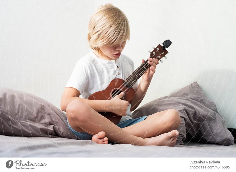 Young boy tuning ukulele at home. Blond haired boy sitting on couch playing acoustic guitar. young cheerful hobby people song music instrument teenager