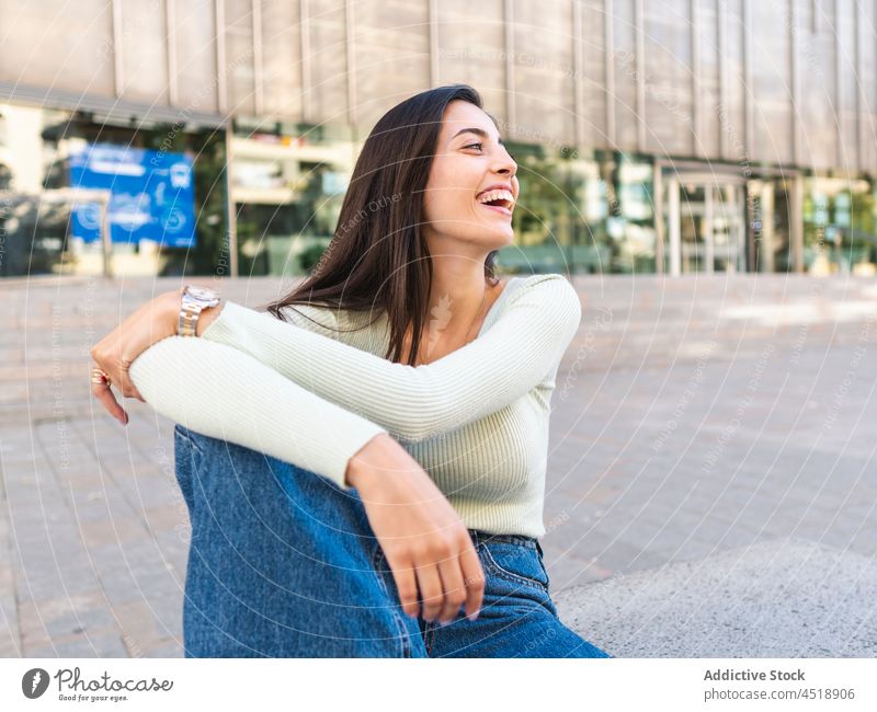 Positive female sitting on stone border on street in daytime woman city urban smile happy embracing knee cheerful style positive charming town joy trendy