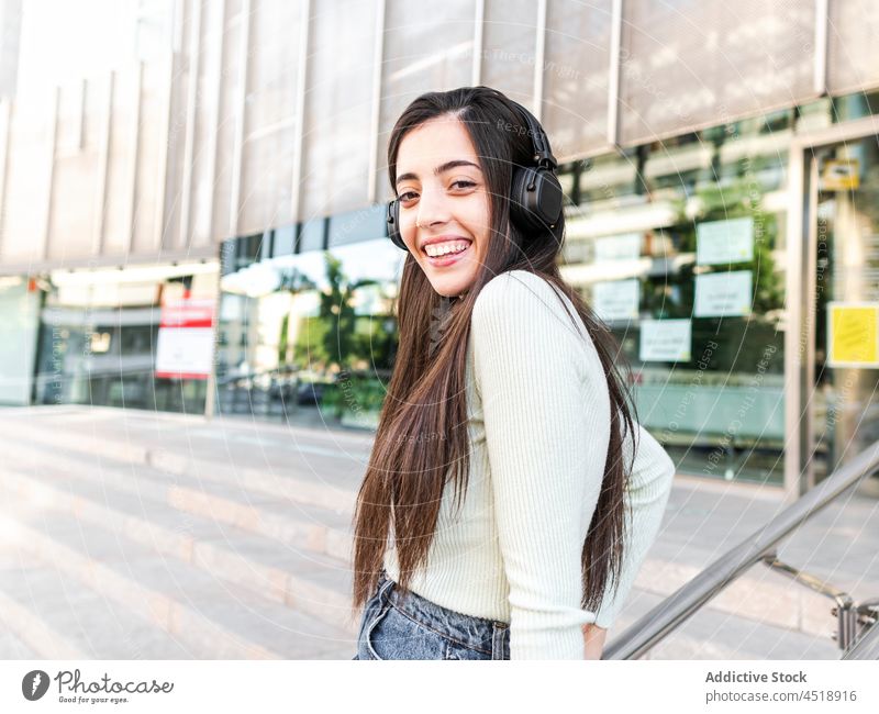 Happy female in headphones standing on street woman dream music calm city happy appearance style urban building adjust brown hair charming outfit lady device