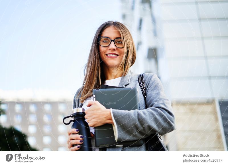 Businesswoman standing with tablet and thermos on street businesswoman entrepreneur style city building device positive trendy female gadget outfit daytime