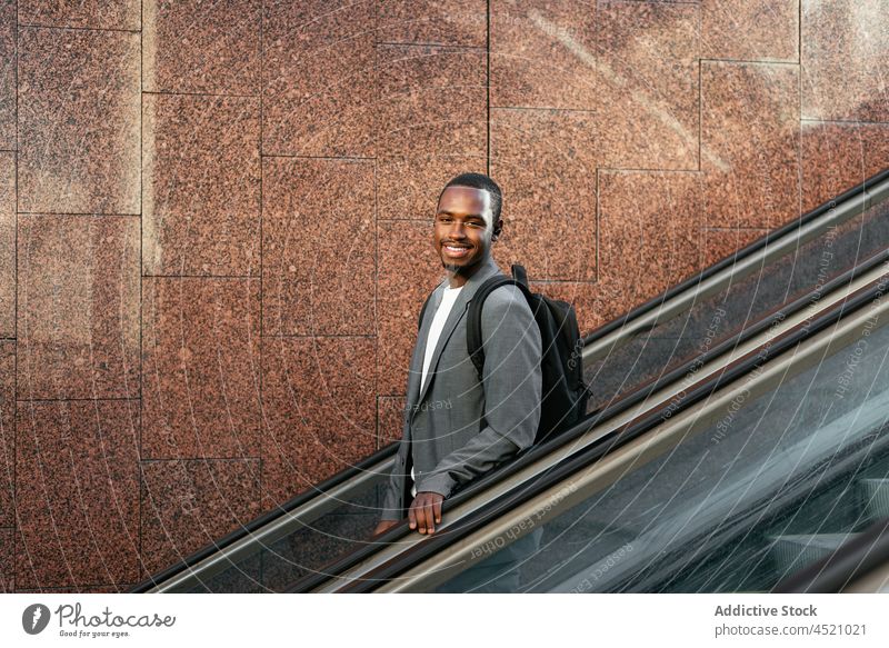 Smiling black man with backpack listening to music in earbuds executive playlist tws song escalator formal male entrepreneur businessman earphones device