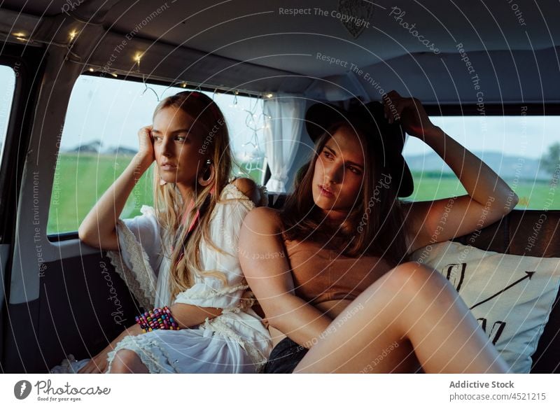 Two beautiful caucasian girls in summer clothes lying on the seat inside a vintage van looking away two pretty cute woman young female brunette blonde tanned
