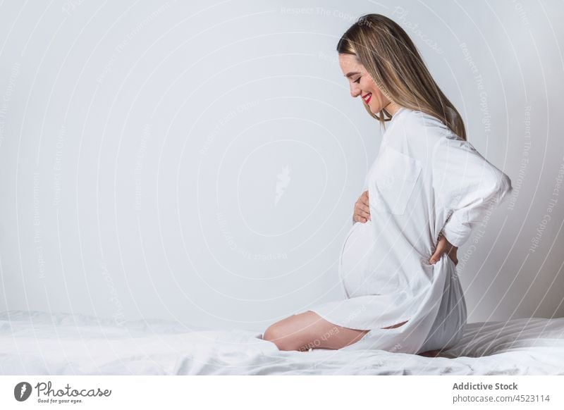 Positive pregnant woman with hand on back sitting on bed belly tender gentle maternal harmony positive anticipate expect female pregnancy smile carefree