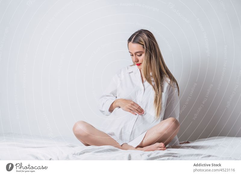 Positive pregnant woman with hand on belly sitting on bed tender gentle maternal harmony positive anticipate expect female pregnancy carefree pleasure bedroom