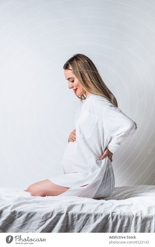 Positive pregnant woman with hand on back sitting on bed belly tender gentle maternal harmony positive anticipate expect female pregnancy smile carefree