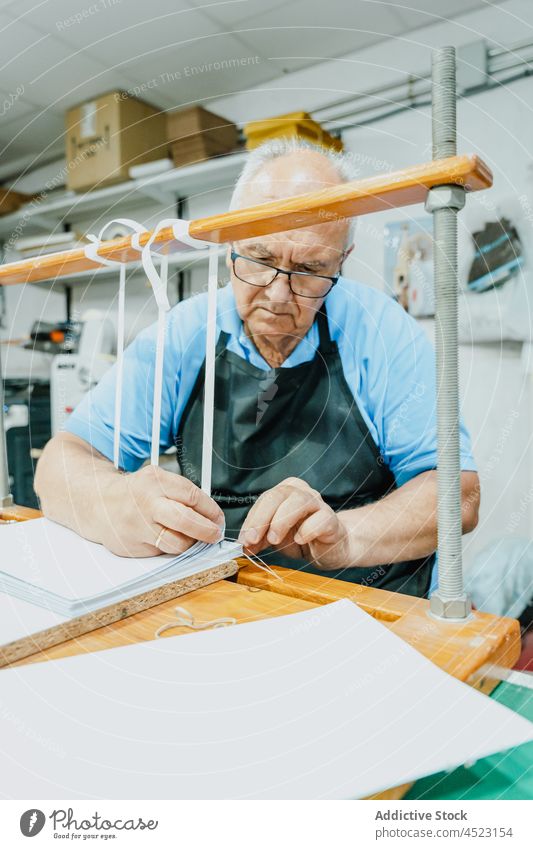 Focused aged male artisan tying tapes on wooden board in printing studio man tie printing machine work attentive process job occupation press senior eyeglasses