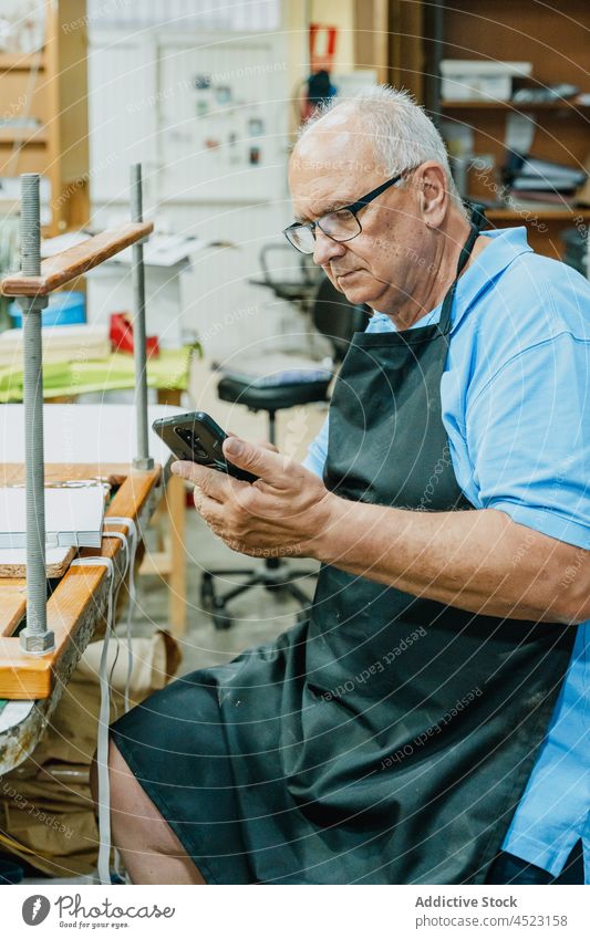 Focused elderly male artisan browsing smartphone in workshop man using print concentrate read workbench mobile senior apron eyeglasses occupation master job