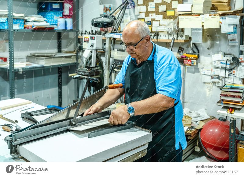 Serious senior male master operating on cutting machine in workshop man carton print artisan process concentrate job serious gray hair eyeglasses apron