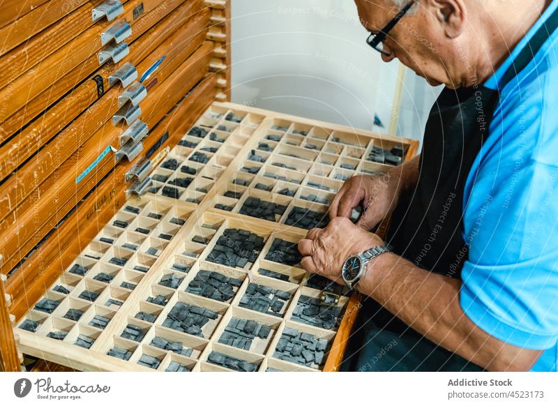 Crop serious elderly male master sitting at type case with lead letters for letterpress man choose workshop print vintage artisan box senior apron eyeglasses