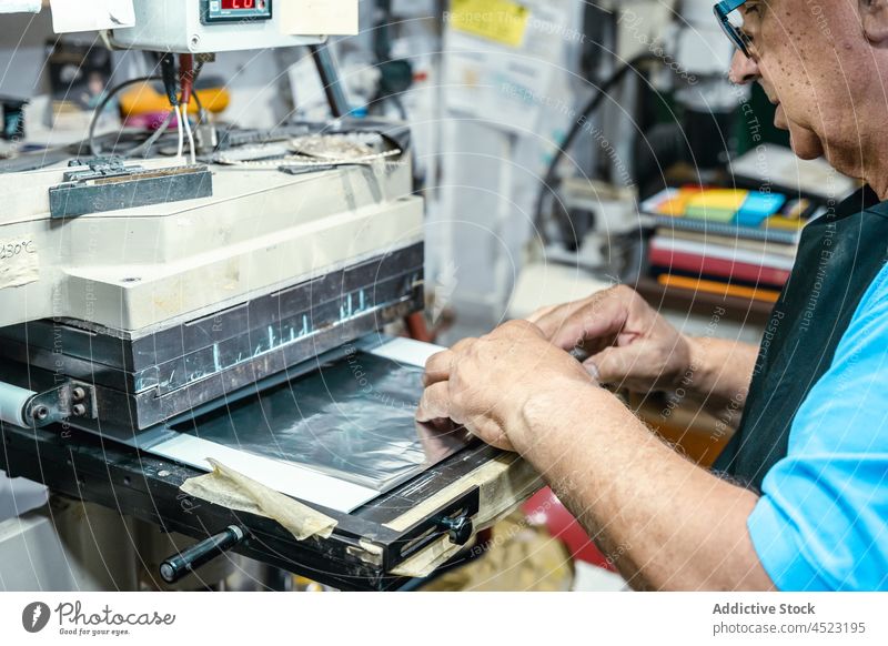 Crop elderly artisan using laminating machine in printing studio man operate lamination printing machine work attentive process job male aged apron eyeglasses