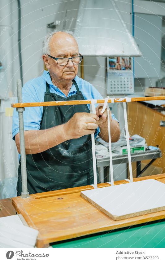 Focused aged male artisan tying tapes on wooden board in printing studio man tie printing machine work attentive process job occupation press senior eyeglasses