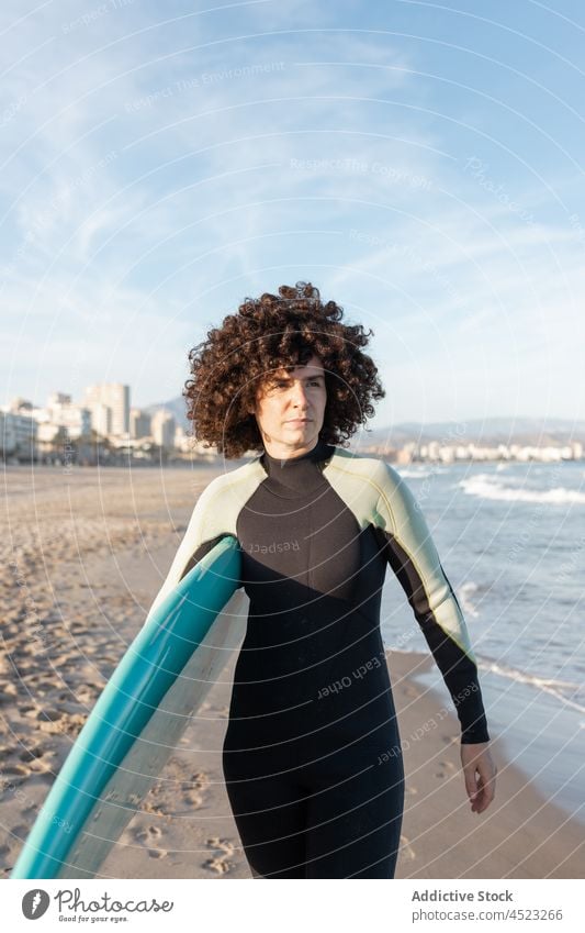 Curly haired woman walking with surfboard on sea shore surfer tropical beach wave foam carefree activity female seashore wetsuit coast water cloudless sky bay