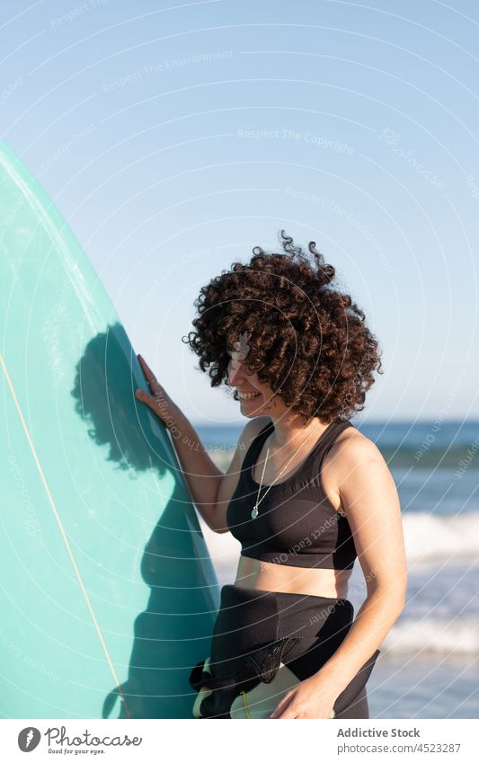 Curly haired woman with surfboard on sea shore surfer smile tropical beach wave foam delight happy carefree activity female seashore wetsuit glad coast water