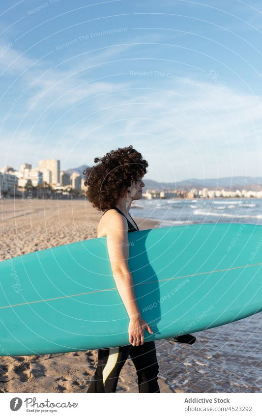 Curly haired woman with surfboard on sea shore surfer tropical beach wave foam carefree activity female seashore wetsuit coast water cloudless sky bay scenery