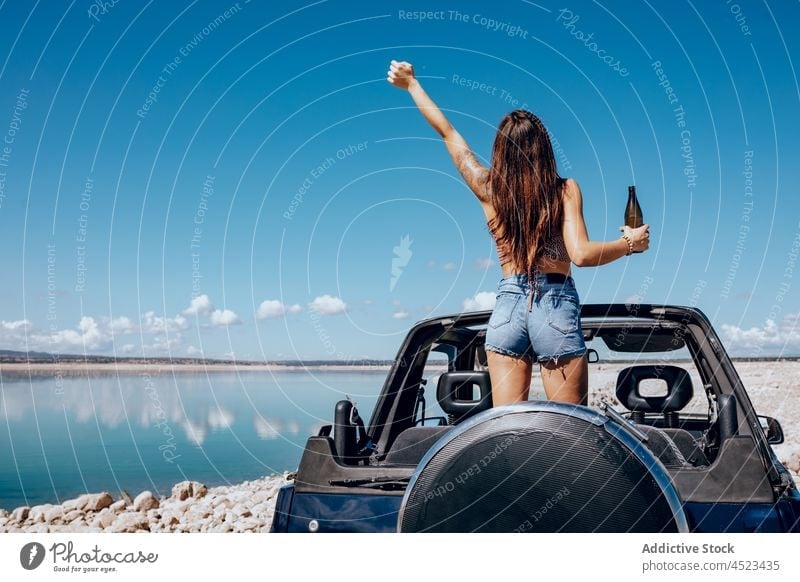 Cheerful woman standing on car with raised bottle of beer safari happy river chill cool freedom female adventure carefree arms raised relax arm raised nature