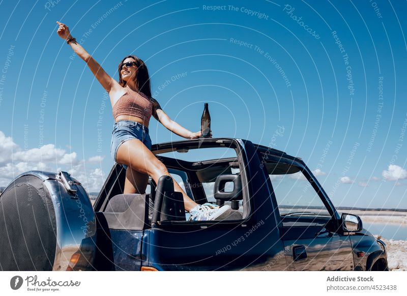 Cheerful woman standing on car with raised bottle of beer safari happy river chill cool freedom female adventure carefree sunglasses relax arm raised nature
