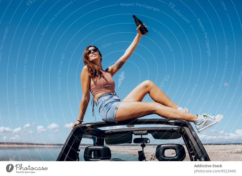 Cheerful woman sitting on car with raised bottle of beer safari happy river chill cool freedom female adventure carefree sunglasses relax arm raised nature roof