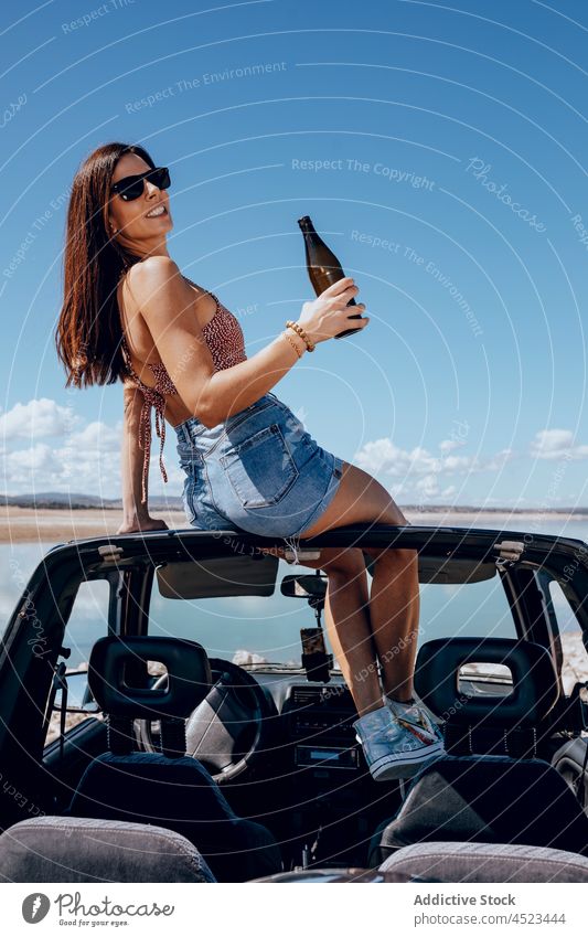 Cheerful woman sitting on car with bottle of beer safari happy river chill cool freedom female adventure carefree sunglasses relax arm raised nature roof enjoy