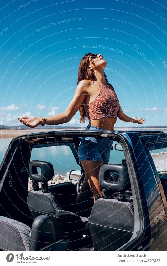 Trendy woman standing in cabriolet and admiring sea admire seascape car convertible road trip traveler beach vacation nature journey female young long hair