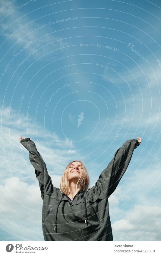 Young blond woman in khaki shirt with hands up in front of blue sky outdoors pretty vertical landscape summer explorer freedom wind blonde hood travel skies