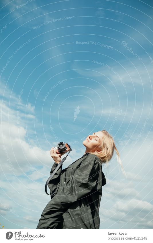 Young blond woman in khaki shirt with a hood holding camera in front of blue sky outdoors photographer eyes closed pretty landscape summer explorer freedom wind