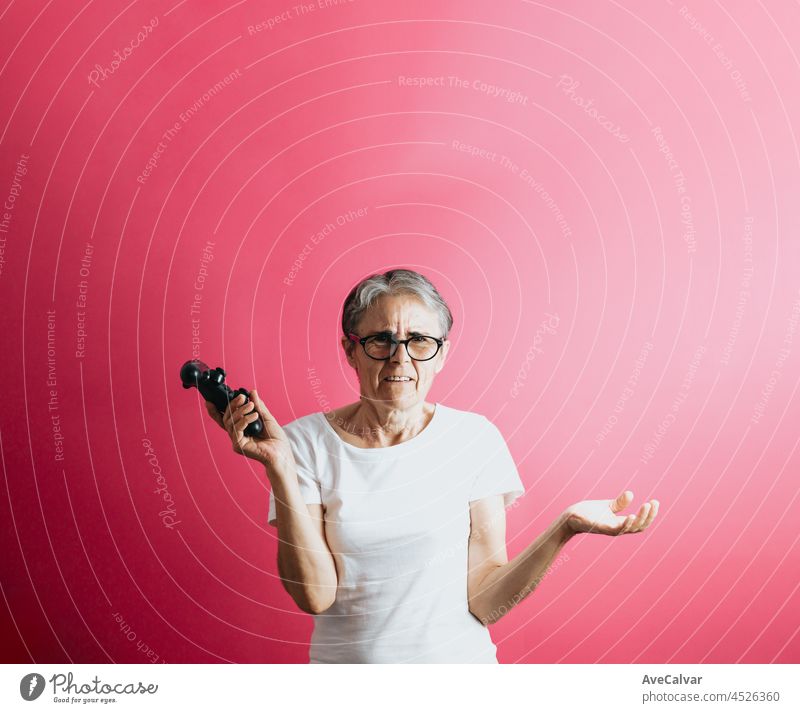 Old senior woman confused while holding a console pad, video games playing pastel pink removable background, video games old people, copy space, white shirt space