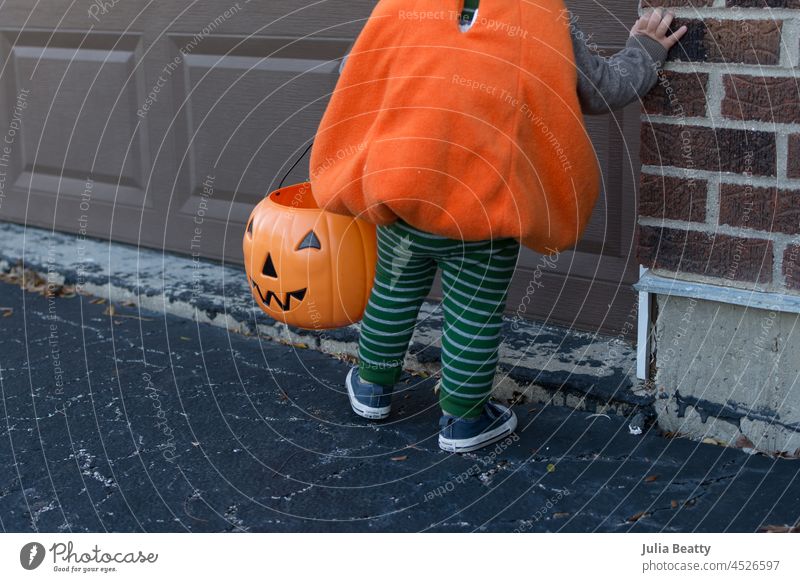 18 month old toddler wearing a jack-o-lantern pumpkin costume while trick or treating; plastic trick or treat bucket halloween orange fleece cozy holiday autumn