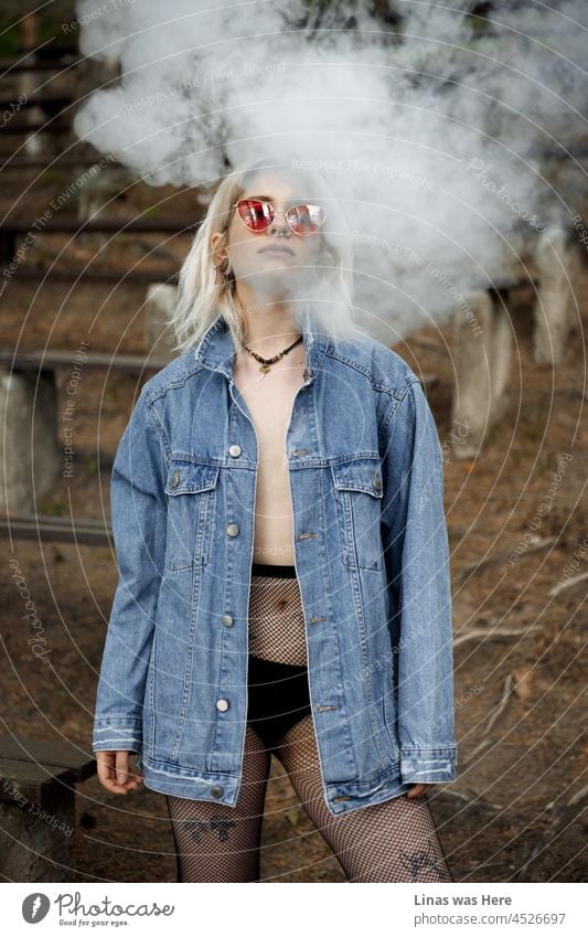 A gorgeous blonde girl dressed in black lingerie, blue jeans jacket, and fashionable shades is being all sexy and wild. A fine portrait of a young model. Smoke in the background goes well with this smoking beauty.