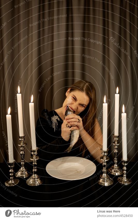 Dinner time. Time for some herring. Gorgeous and wild brunette girl biting off the fish head without any cutlery. Dressed in a fancy black dress, surrounded by candles, and fine curtains behind her. Some shiny jewelry compliments her as well.