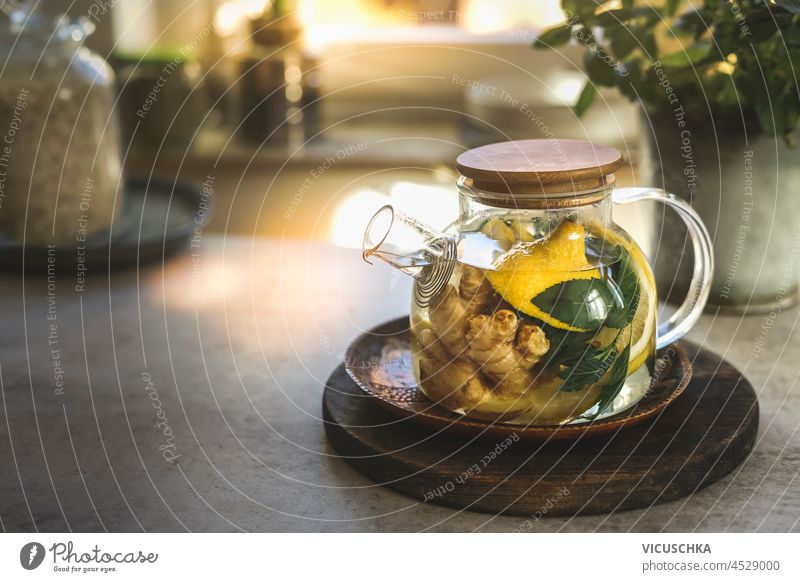 Glass tea pot with mint leaves, lemon and ginger on kitchen table with utensils and window background. Herbal tea with healthy ingredients and vitamin c in cold season. Natural treatment. Front view.