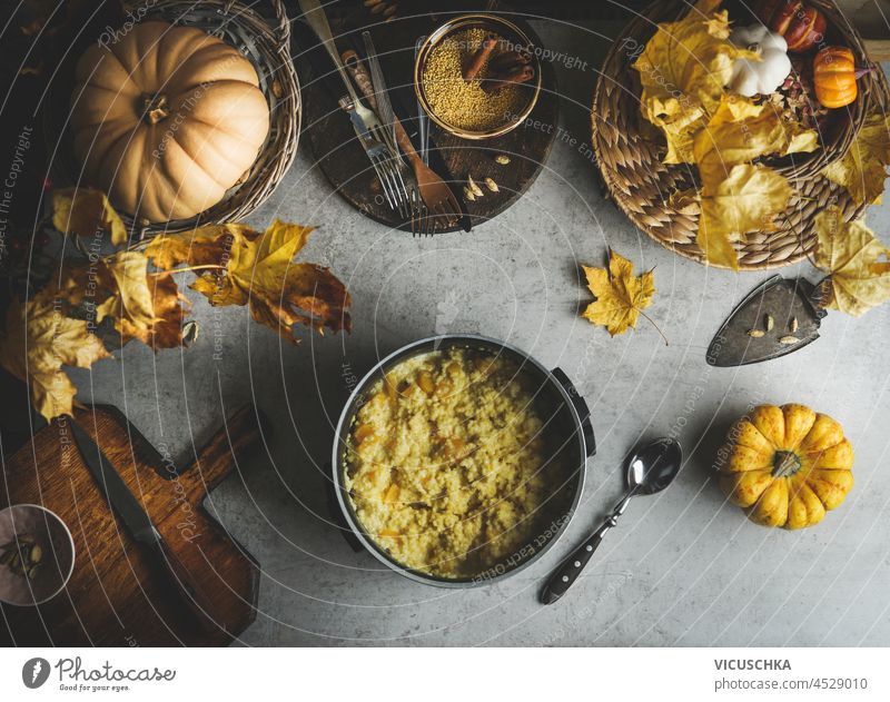 Millet porridge with pumpkins in cooking pot on grey concrete table with yellow autumn leaves, wooden cutting board, knife, spoon and kitchen utensils. Cooking healthy vegan breakfast at home.Top view
