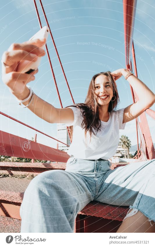 Young urban mixed race woman taking a selfie while smiling during a sunny day, urban lifestyle, white tshirt blank space, trendy social network, young people, wide angle image